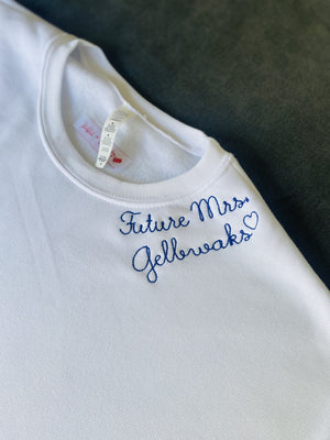 A close-up of a white crewneck sweatshirt featuring the phrase 'Future Mrs. Jellows' embroidered in blue cursive script with a small heart, laid flat on a dark textured surface.