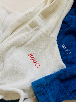 Close-up of plush bathrobes in cream and blue, each embroidered with a small logo. The cream robe is in the foreground with red embroidery reading 'juju' and a smile, and the blue robe behind it with white embroidery reading 'ari' and a smile.