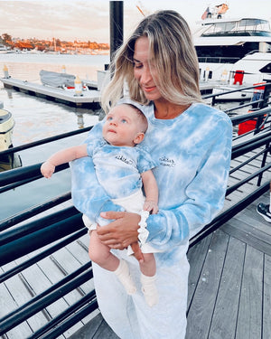 Woman holding a baby both wearing blue tie-dye outfits by a marina.