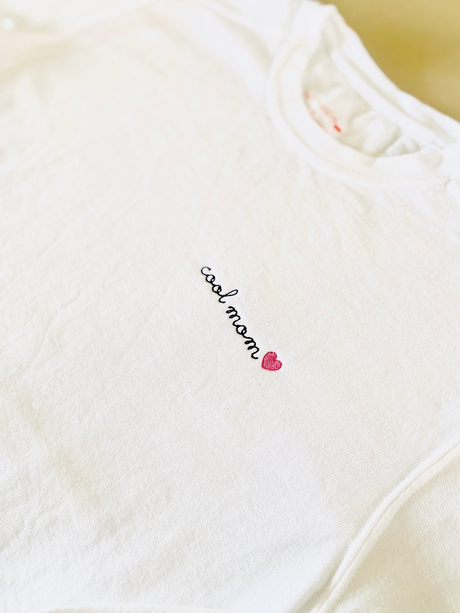 White crewneck sweatshirt with 'cool mom' text and a heart symbol embroidered on the left chest area.